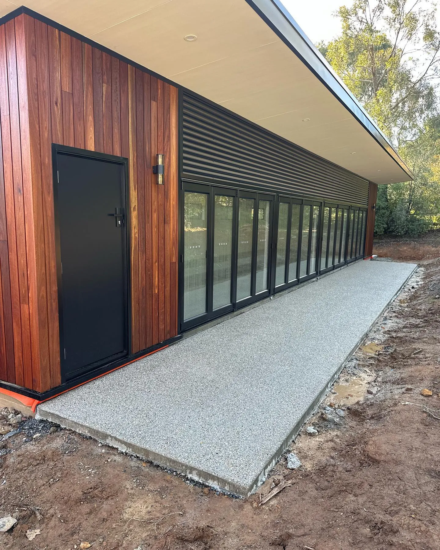 Light exposed-aggregate path running the full length of a timber-clad architectural studio with louvre walls
