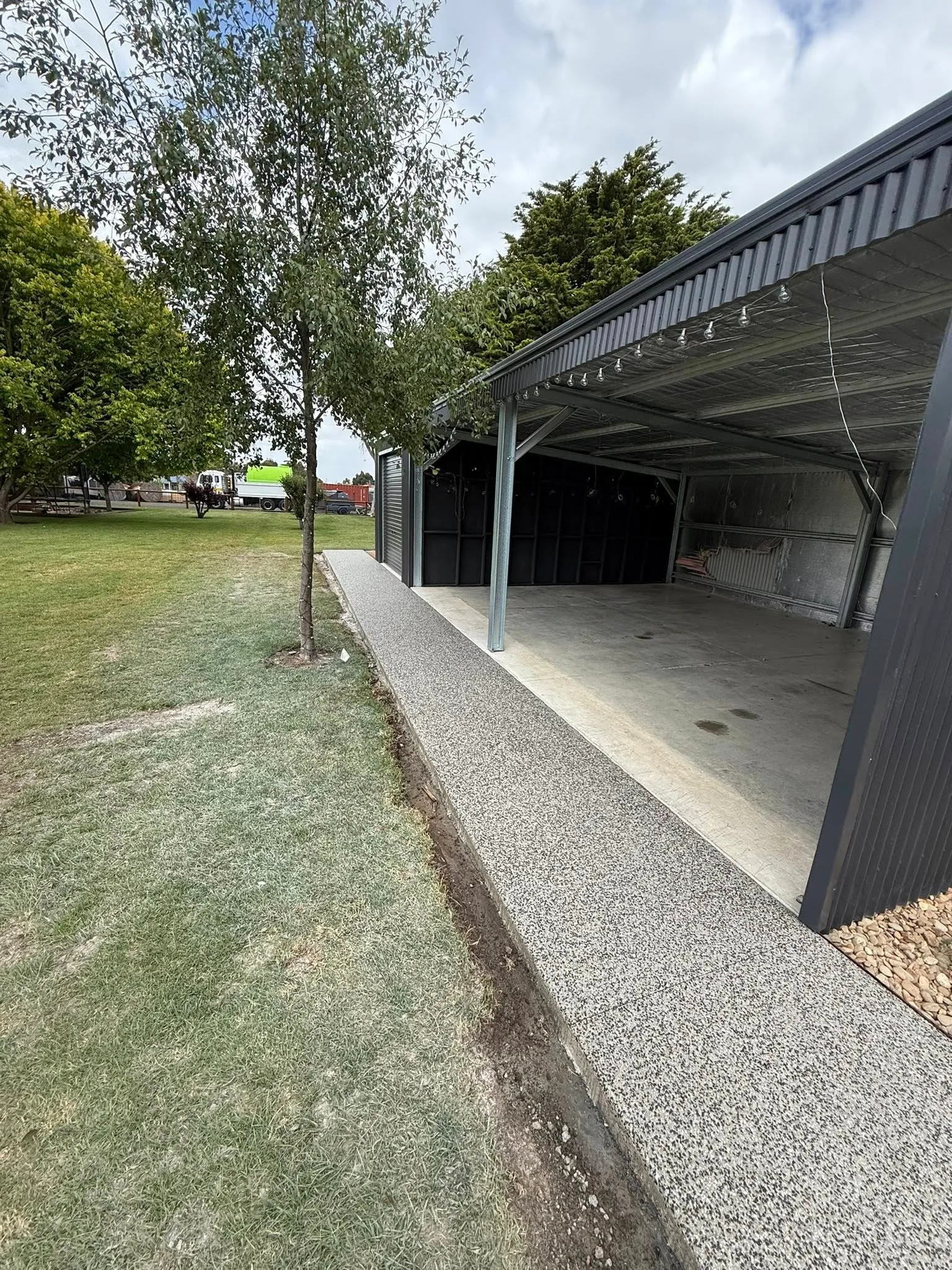 Exposed aggregate strip running alongside a long open-bay shed with mature trees