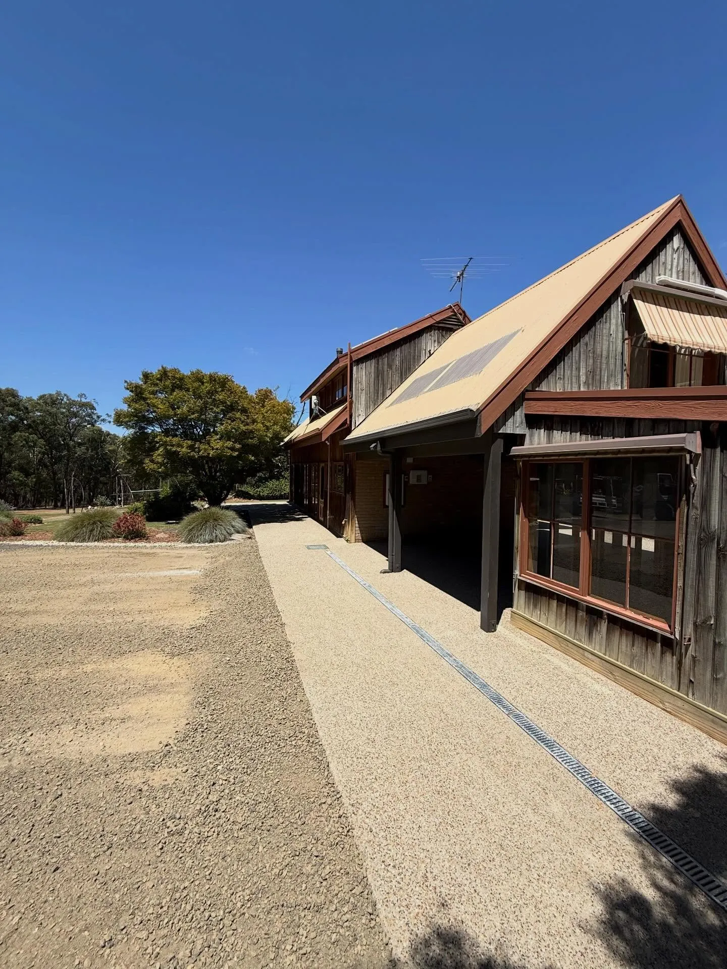 Exposed aggregate path running along a timber barn-style rural home with drainage strip, sharp blue-sky day