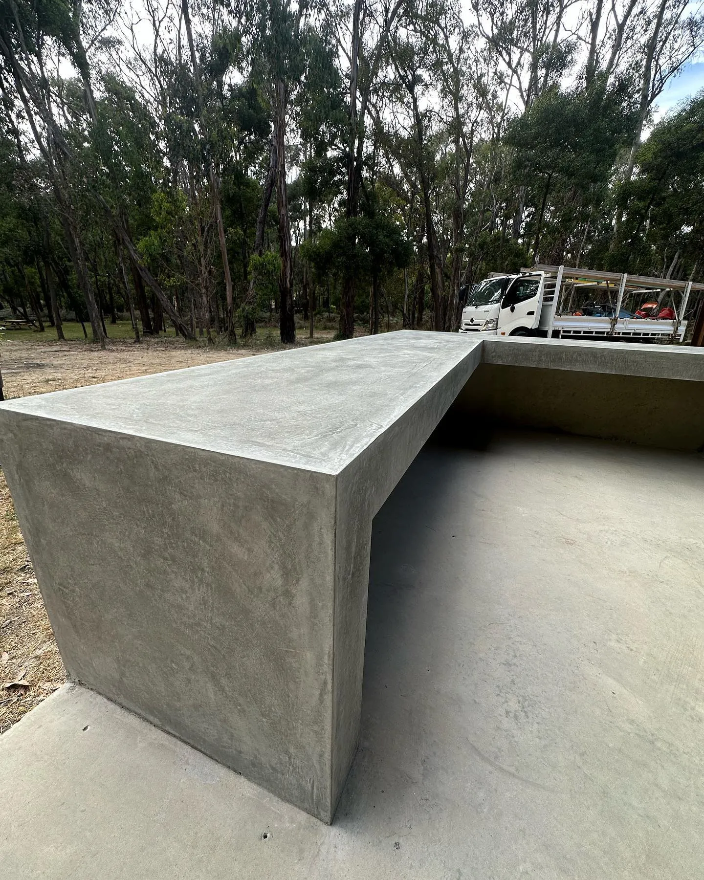 Monolithic raw concrete bench in a bush clearing with BMT truck parked beyond, sculptural off-form finish