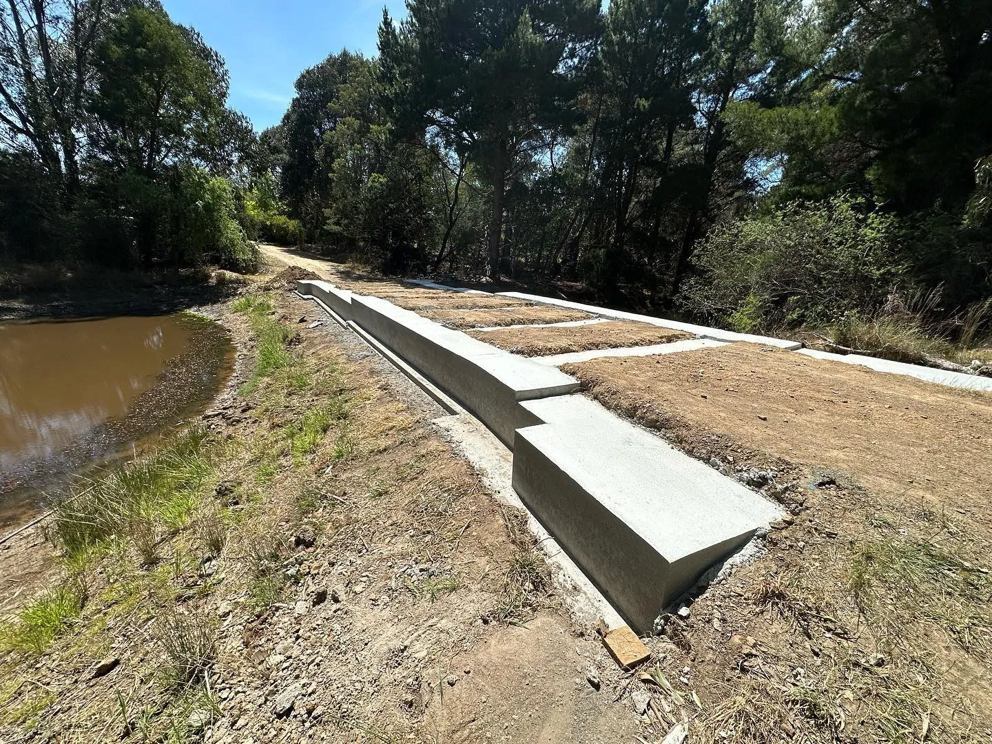 Finished concrete dam-crossing causeway with stepped walls beside still water and surrounding bush