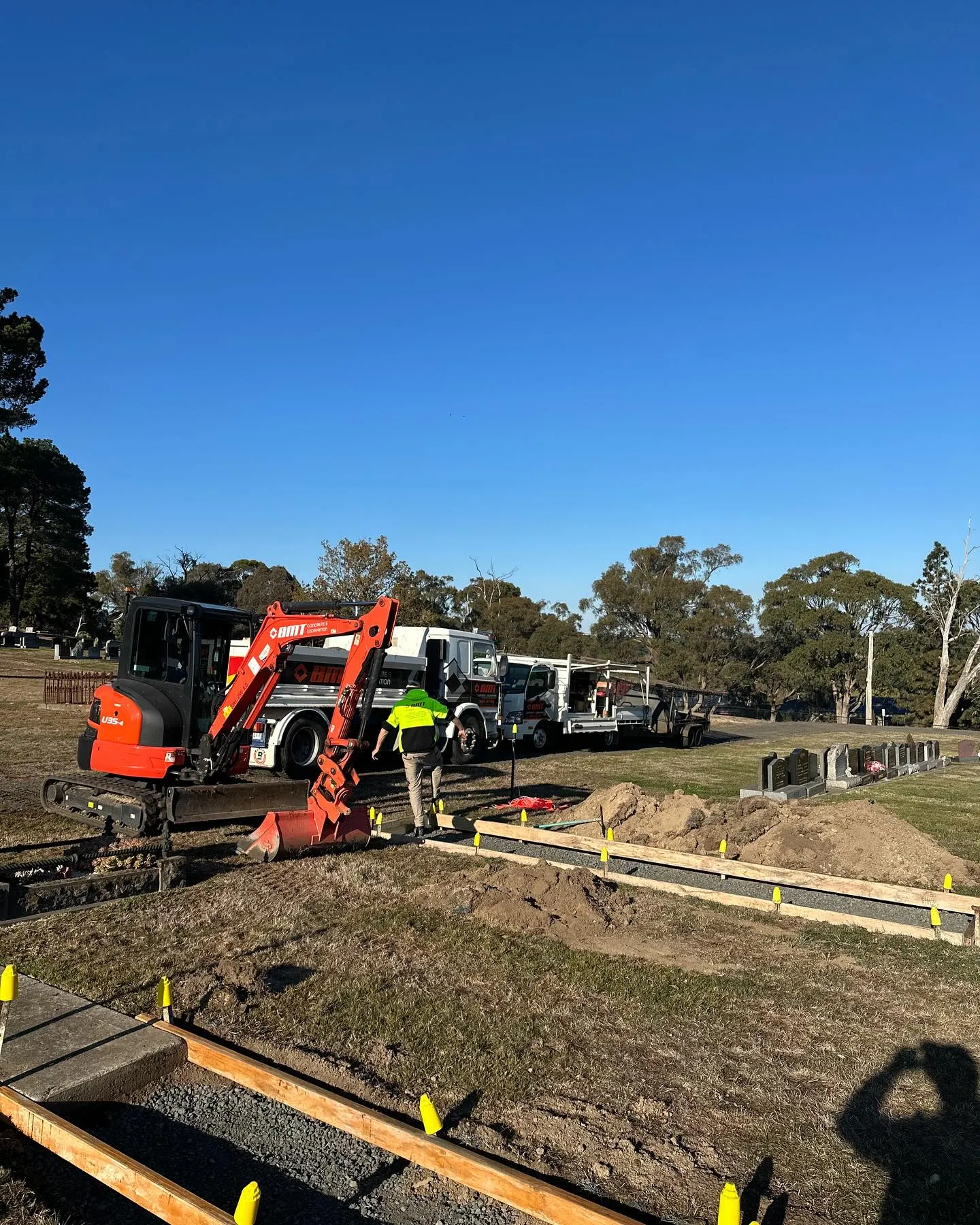 BMT skid-steer, branded truck and a crew member preparing cemetery row formwork under a bright blue sky