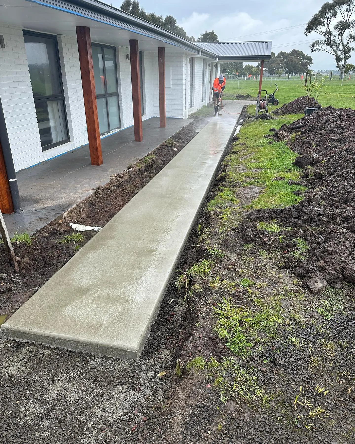 Long narrow concrete path running beside a white rural home, freshly poured with a wet edge