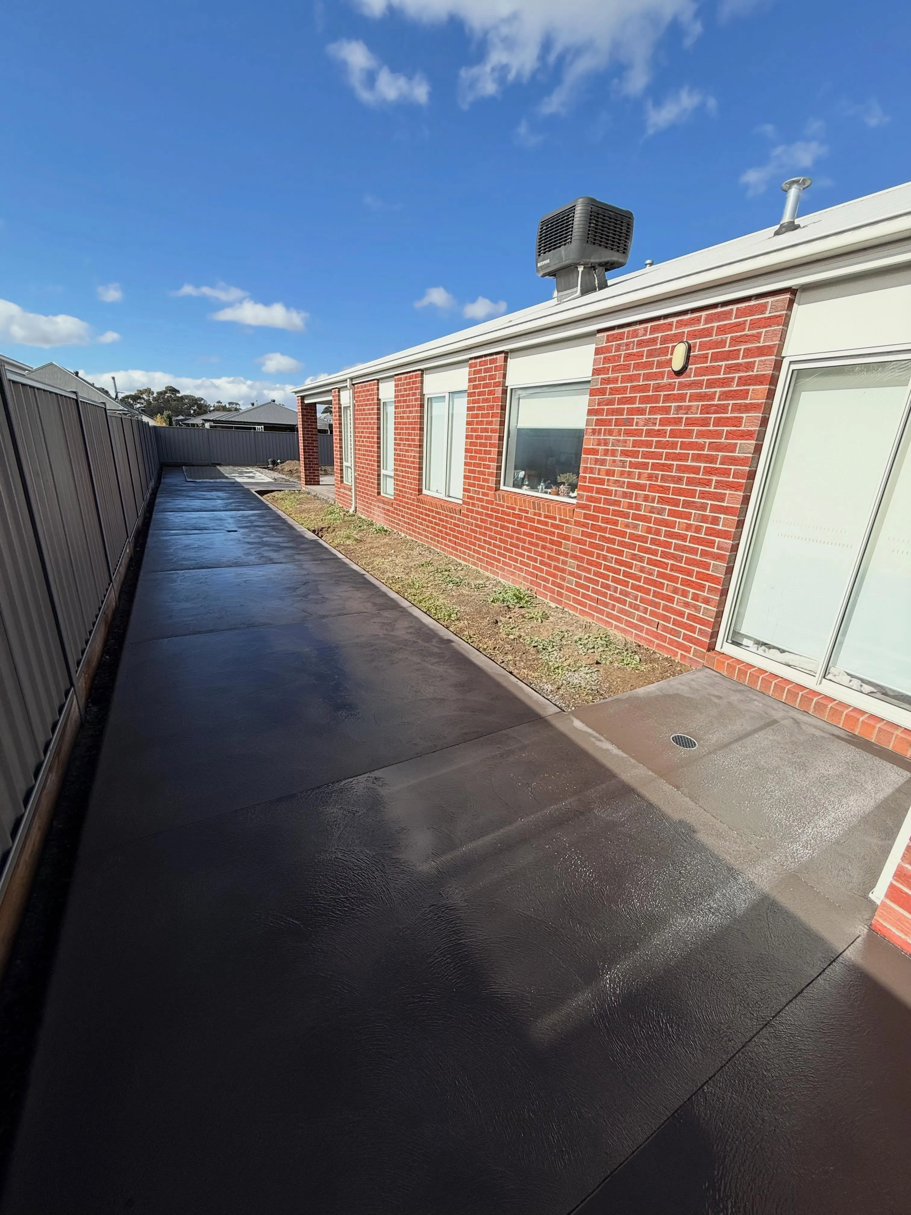 Long wet-look sealed concrete path beside a red-brick rural home running to the fence line